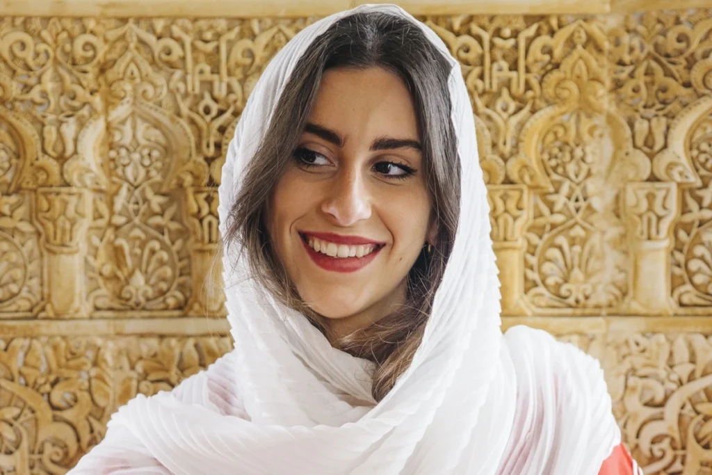 Mulher sorridente usando véu branco em frente a parede decorada com arabescos dourados.