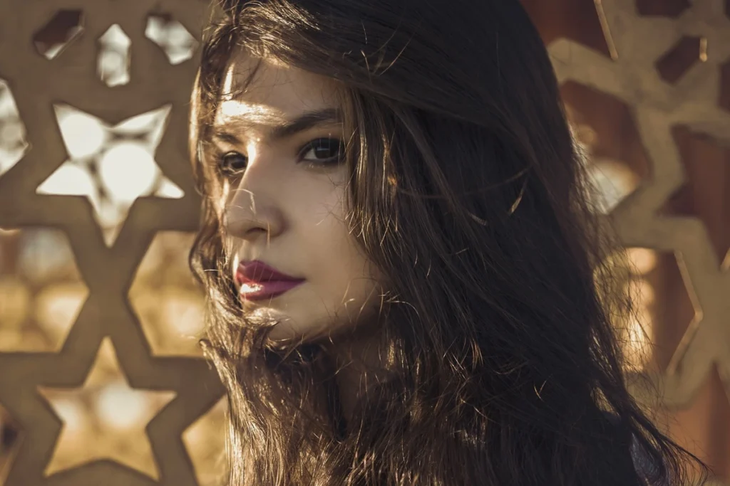 Mulher com cabelos longos ao vento olhando para o lado, iluminada pelo sol, em frente a padrão geométrico árabe.