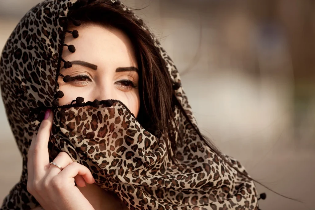 Mulher cobrindo parte do rosto com lenço de estampa de oncinha, mostrando apenas os olhos e sobrancelhas marcadas.