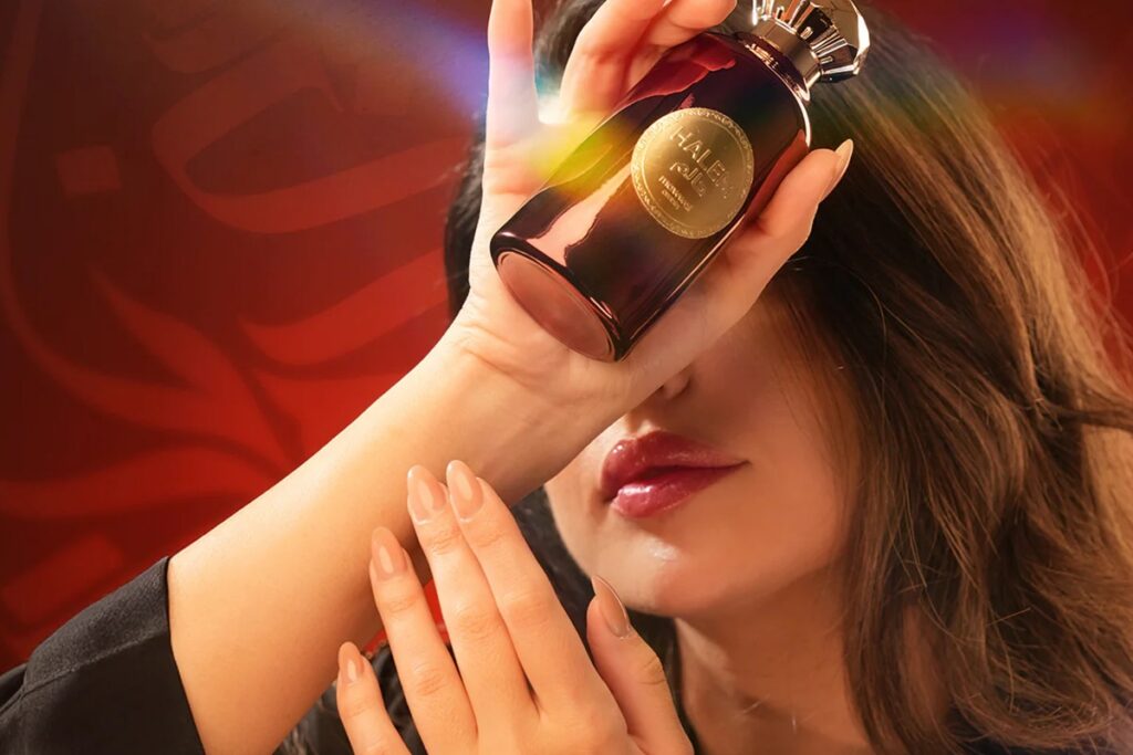 Mulher aplicando perfume árabe premium no pulso, segurando um frasco dourado e vermelho em um ambiente sofisticado.
