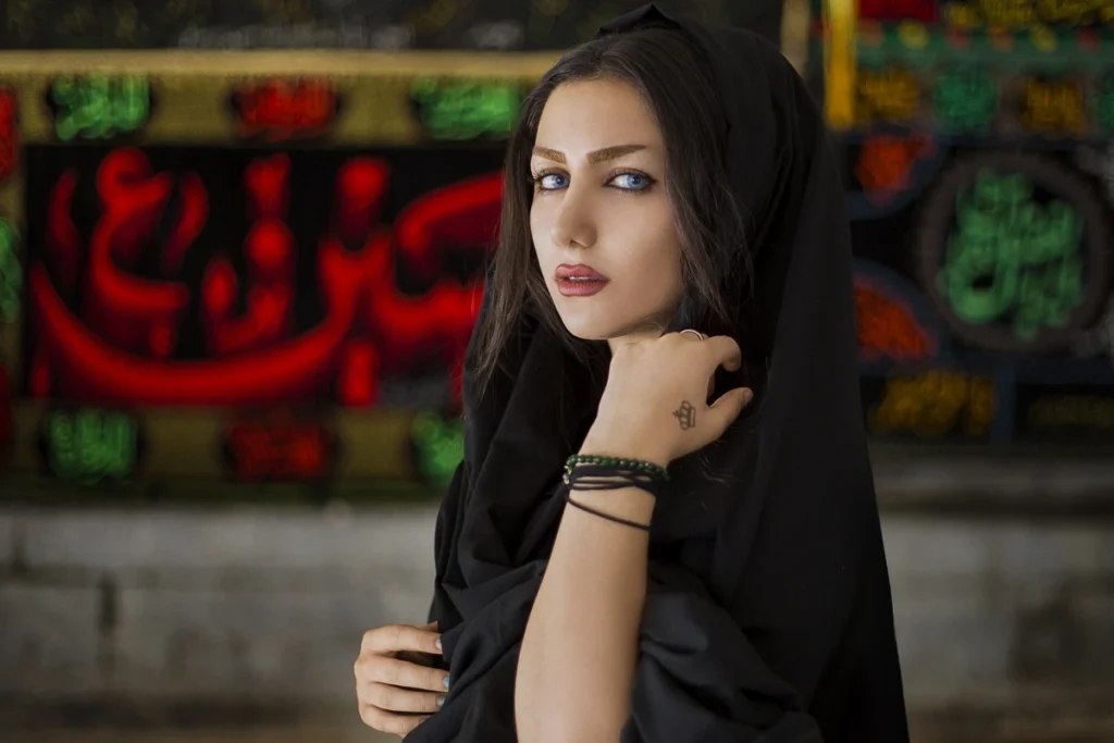 Mulher usando hijab preto com olhar intenso em frente a fundo decorado com caligrafia árabe colorida.