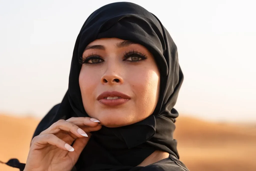 Mulher usando hijab preto olhando para a câmera no deserto, com maquiagem destacada e luz suave.