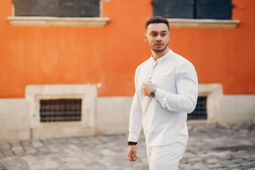 Homem elegante caminhando em rua de paralelepípedos, usando camisa branca e relógio prateado, em frente a parede laranja.