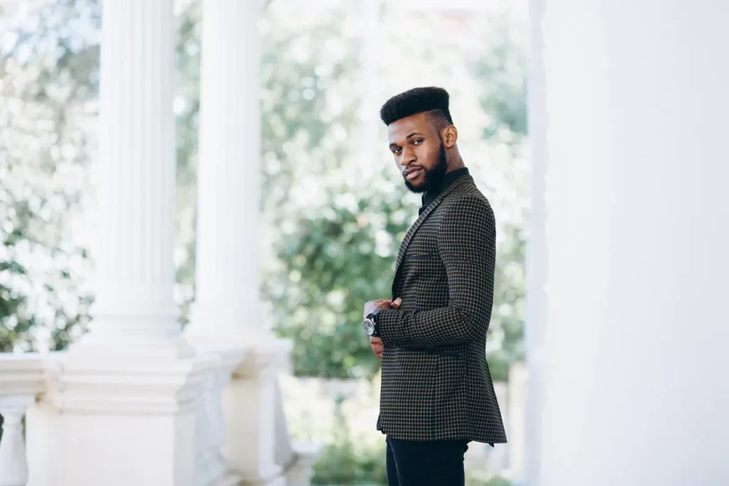 Homem negro com barba e cabelo estilizado vestindo terno xadrez escuro em ambiente externo com colunas brancas e fundo verde