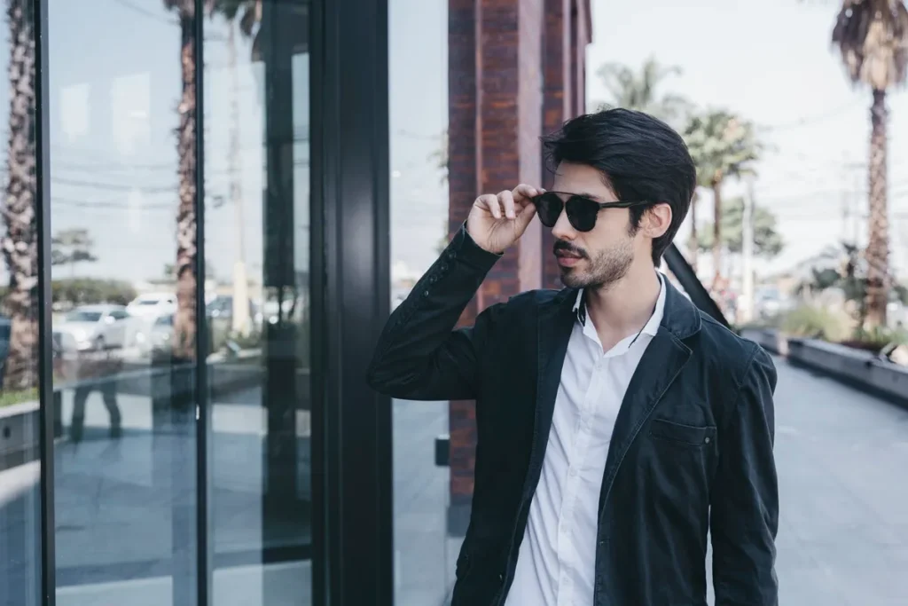 Homem de cabelo escuro usando óculos de sol e blazer preto sobre camisa branca em ambiente urbano com palmeiras ao fundo