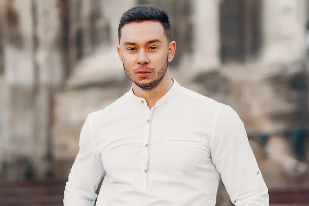 Homem com barba aparada vestindo camisa branca de botão, em ambiente urbano desfocado ao fundo