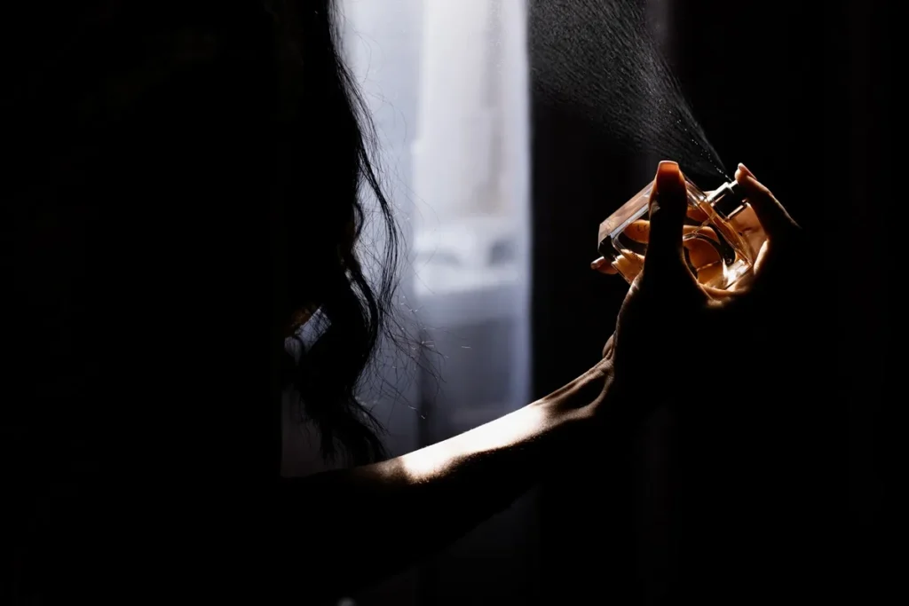 Mão segurando frasco de perfume e borrifando a fragrância em um ambiente escuro com luz suave ao fundo.