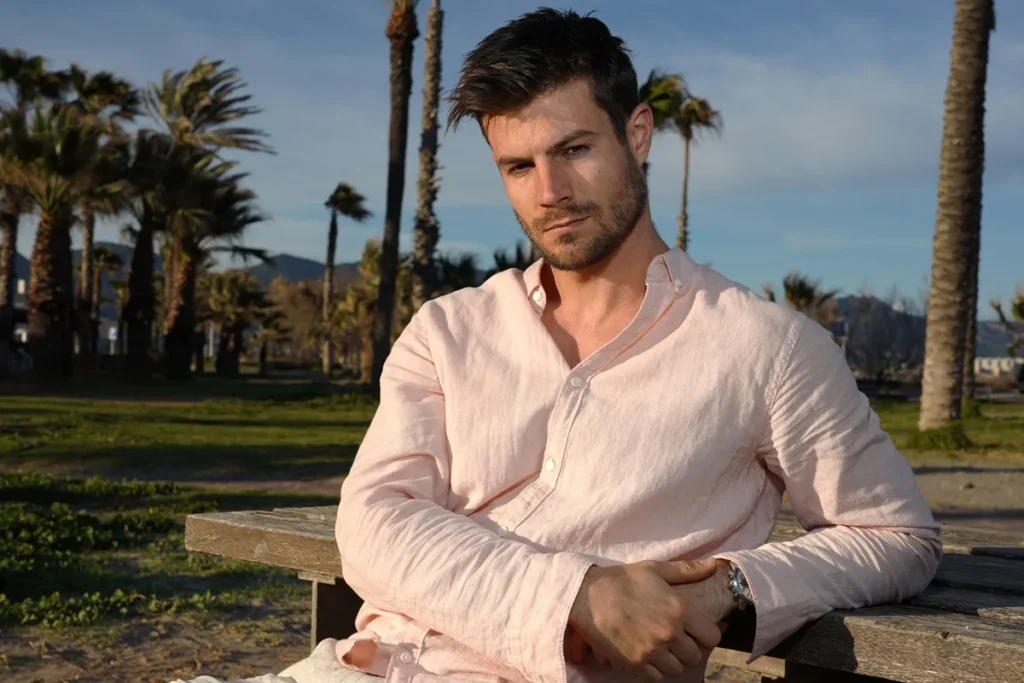 Homem sentado em banco de madeira usando camisa rosa claro, em cenário de praia com palmeiras ao fundo.