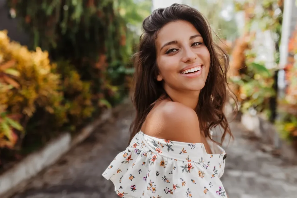 Mulher sorrindo ao ar livre, usando vestido floral ombro a ombro, em ambiente com vegetação colorida.