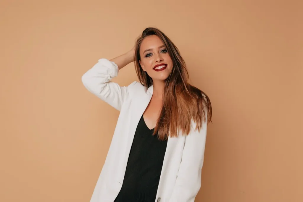 Mulher sorridente usando blazer branco sobre blusa preta, posando em frente a um fundo bege.