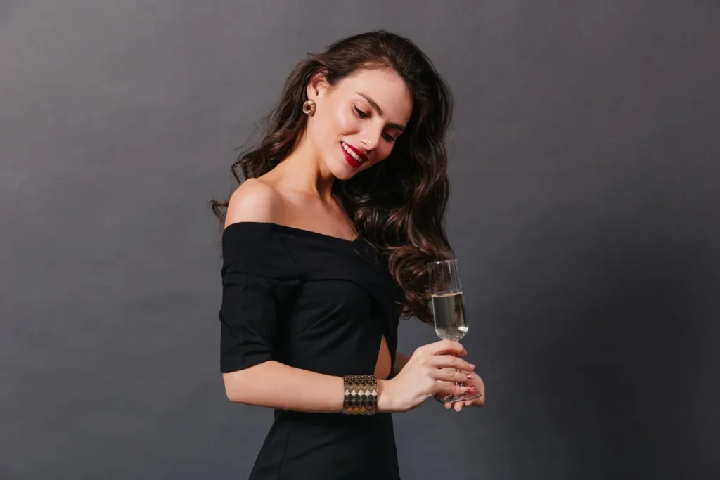 Mulher elegante usando vestido preto e segurando uma taça de champanhe, sorrindo em frente a um fundo cinza.