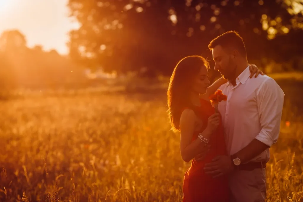 Casal abraçado em um campo durante o pôr do sol, com a mulher segurando uma flor e ambos iluminados pela luz dourada.
