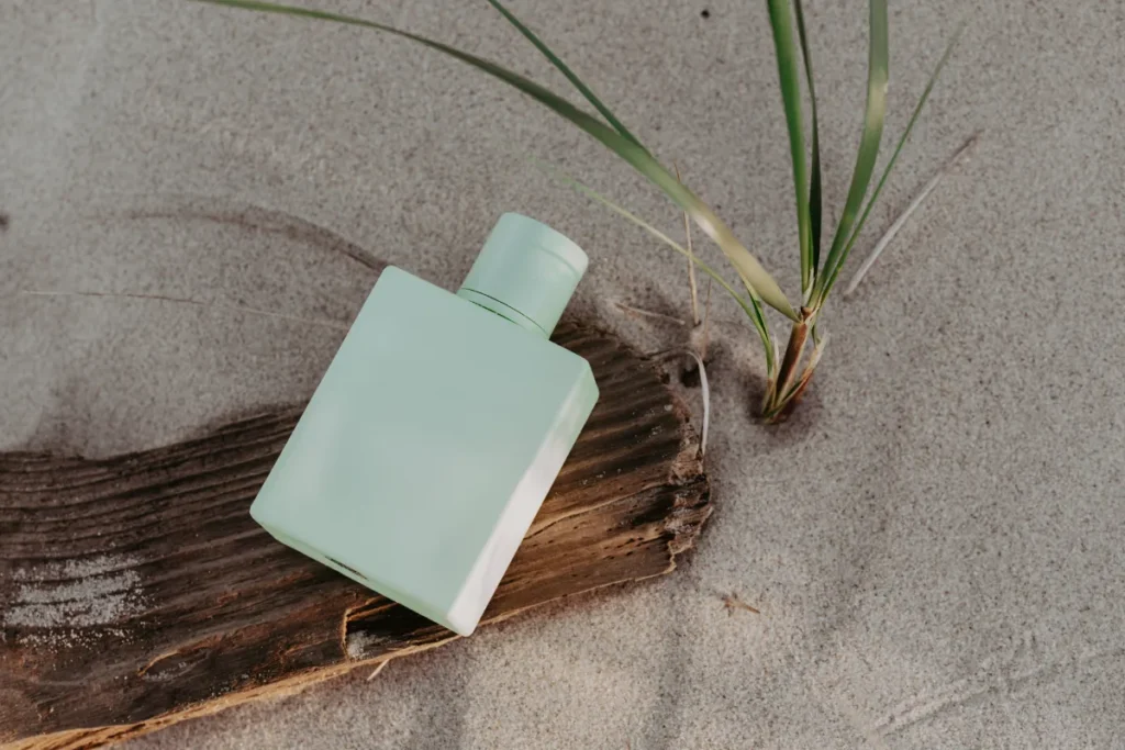 frasco de perfume verde claro colocado sobre areia ao lado de uma pequena planta