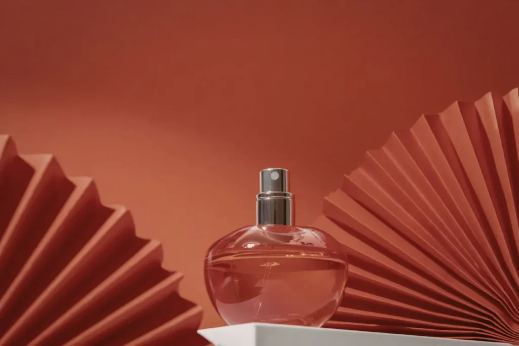 frasco de perfume redondo com líquido rosado colocado sobre base clara com fundo vermelho decorativo