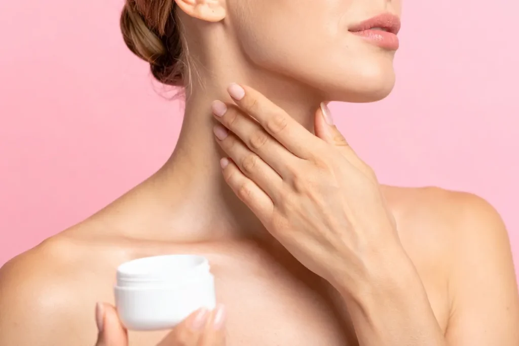 Mulher aplicando creme hidratante no pescoço enquanto segura um pote de creme para cuidados com a pele.
