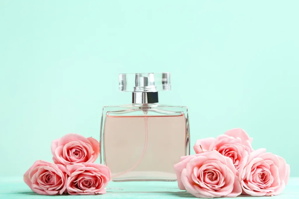 Frasco de perfume transparente com líquido rosado posicionado entre rosas cor-de-rosa sobre fundo verde claro.