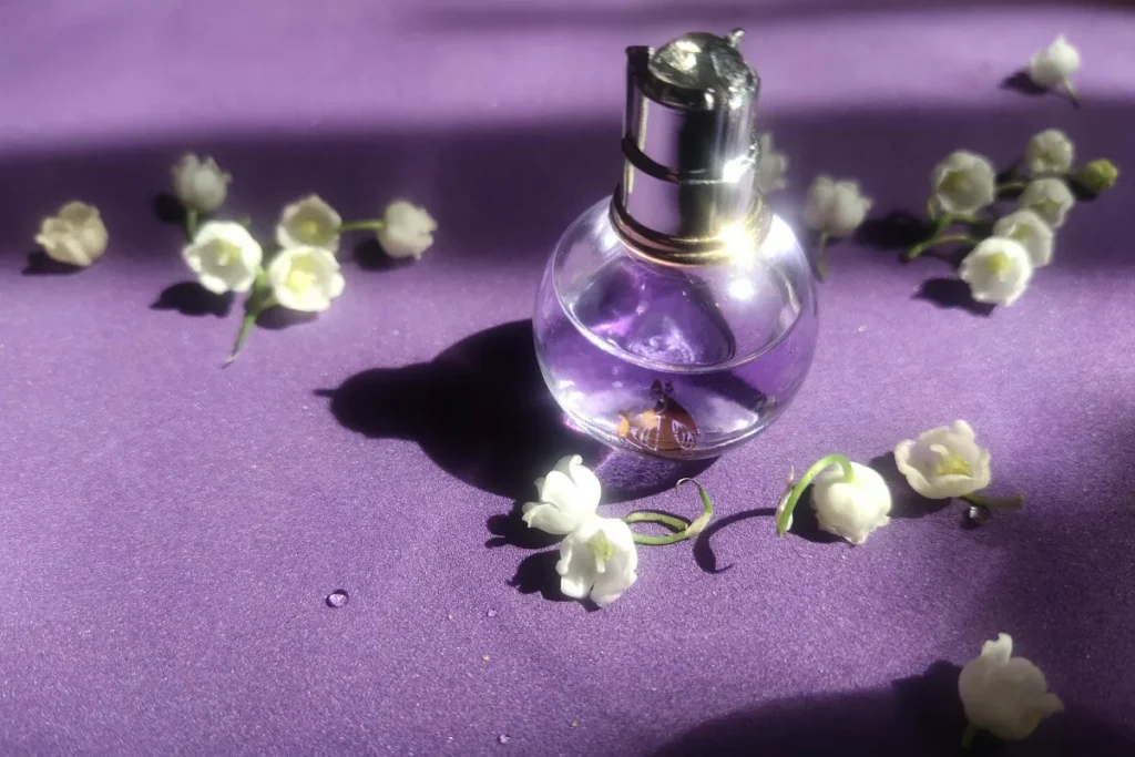 Frasco pequeno de perfume de vidro transparente sobre superfície roxa, cercado por pequenas flores brancas.