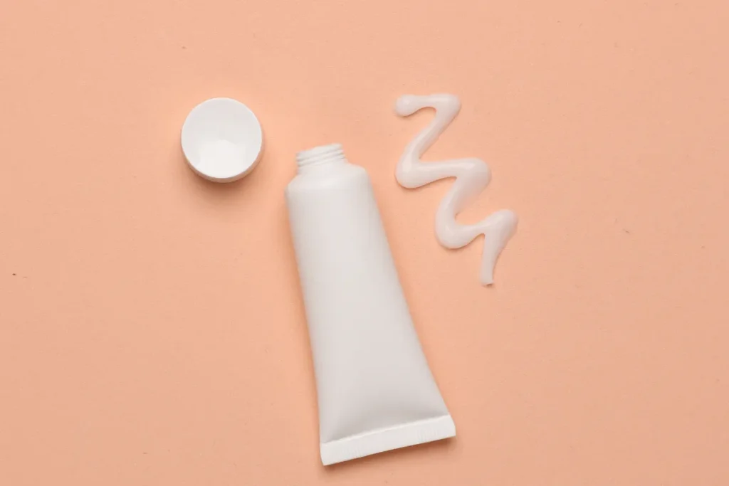 Bisnaga branca de creme hidratante com tampa ao lado e uma pequena quantidade de produto sobre fundo laranja claro.