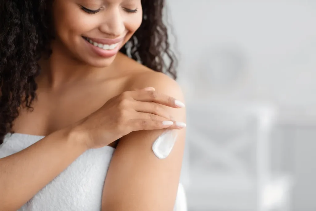 Mulher sorrindo aplica creme hidratante no braço com a mão, em ambiente claro