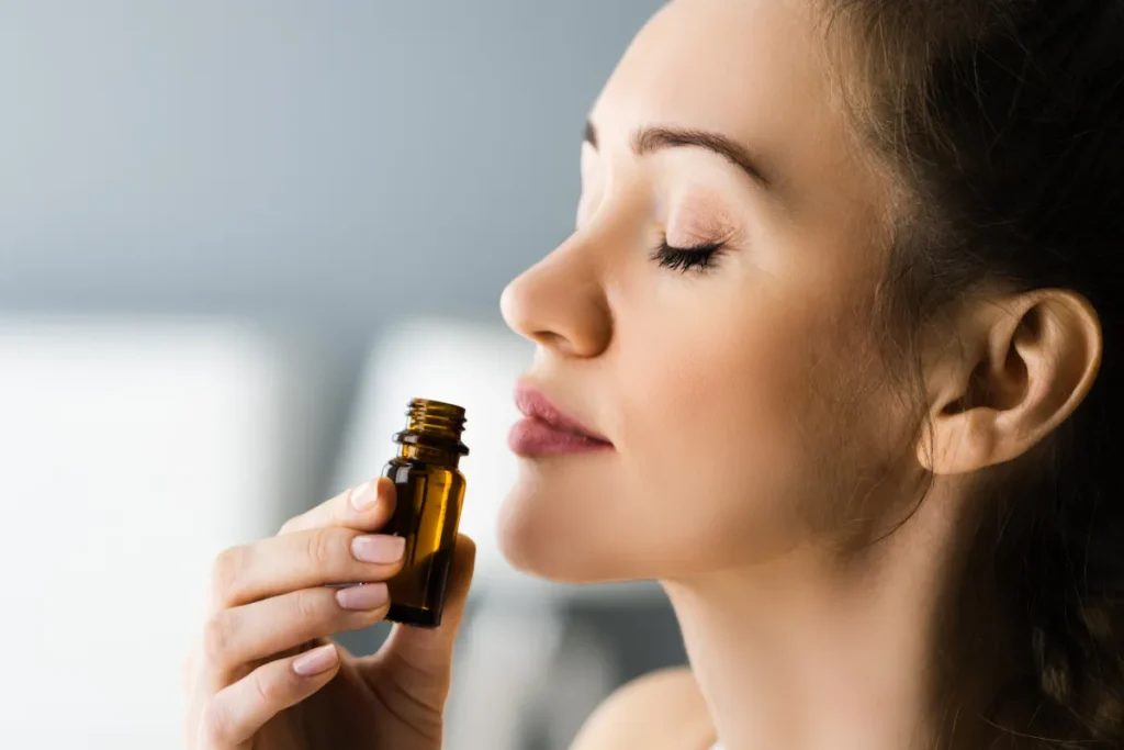 Mulher de olhos fechados aproximando um pequeno frasco âmbar do nariz para sentir o aroma.