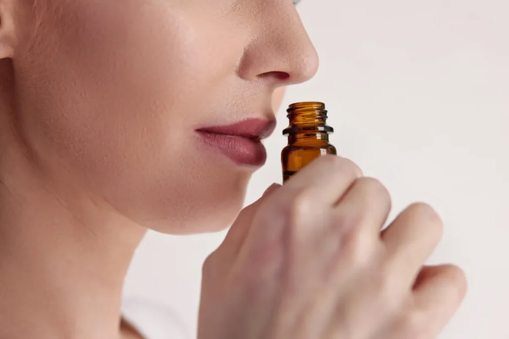 Close no rosto de uma mulher aproximando um pequeno frasco de essência do nariz para sentir o aroma.