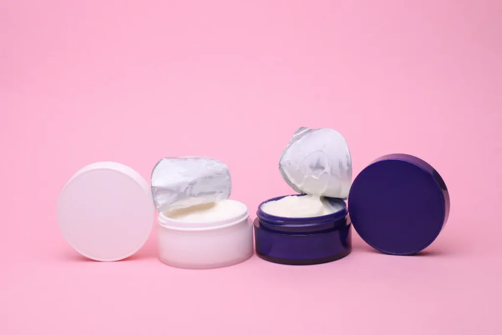 Dois potes de creme corporal abertos com o produto visível sobre fundo rosa.