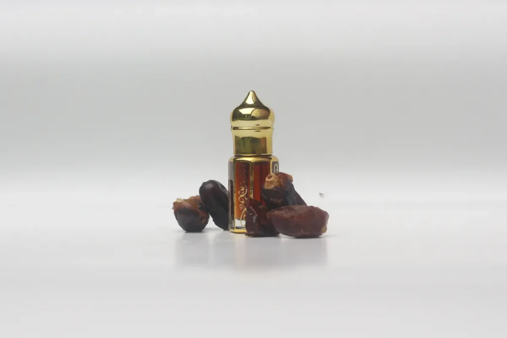 Pequeno frasco de perfume árabe com tampa dourada ao lado de tâmaras sobre fundo branco.