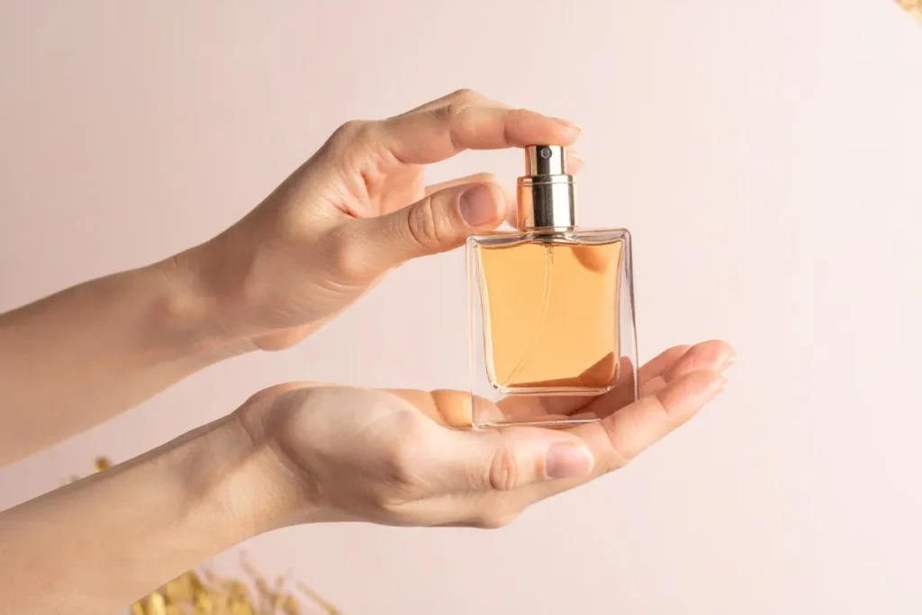 Mãos segurando um frasco de perfume com líquido âmbar enquanto pressionam o borrifador.