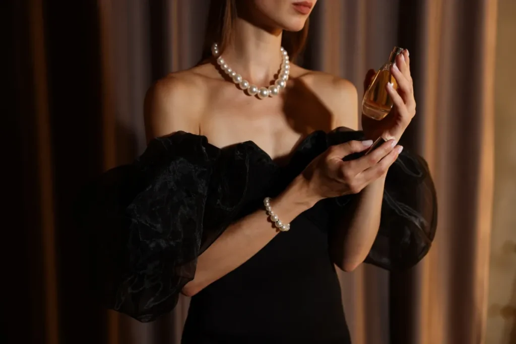 Mulher elegante usando vestido preto e colar de pérolas segurando um frasco de perfume enquanto aplica a fragrância.