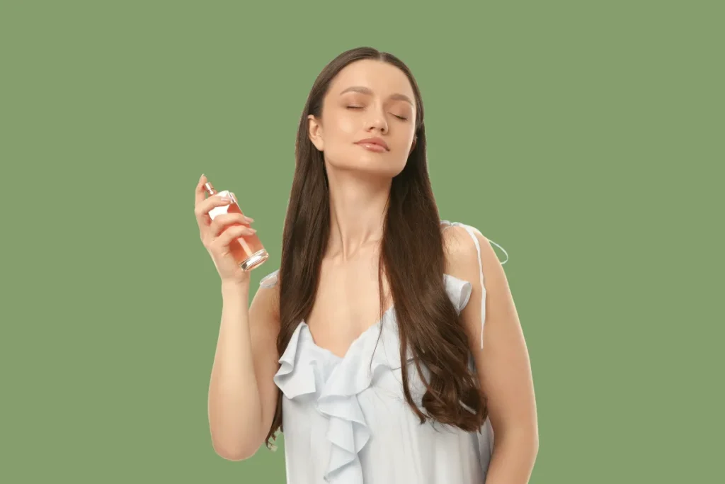 mulher aplicando perfume no pulso com expressão de satisfação em fundo neutro